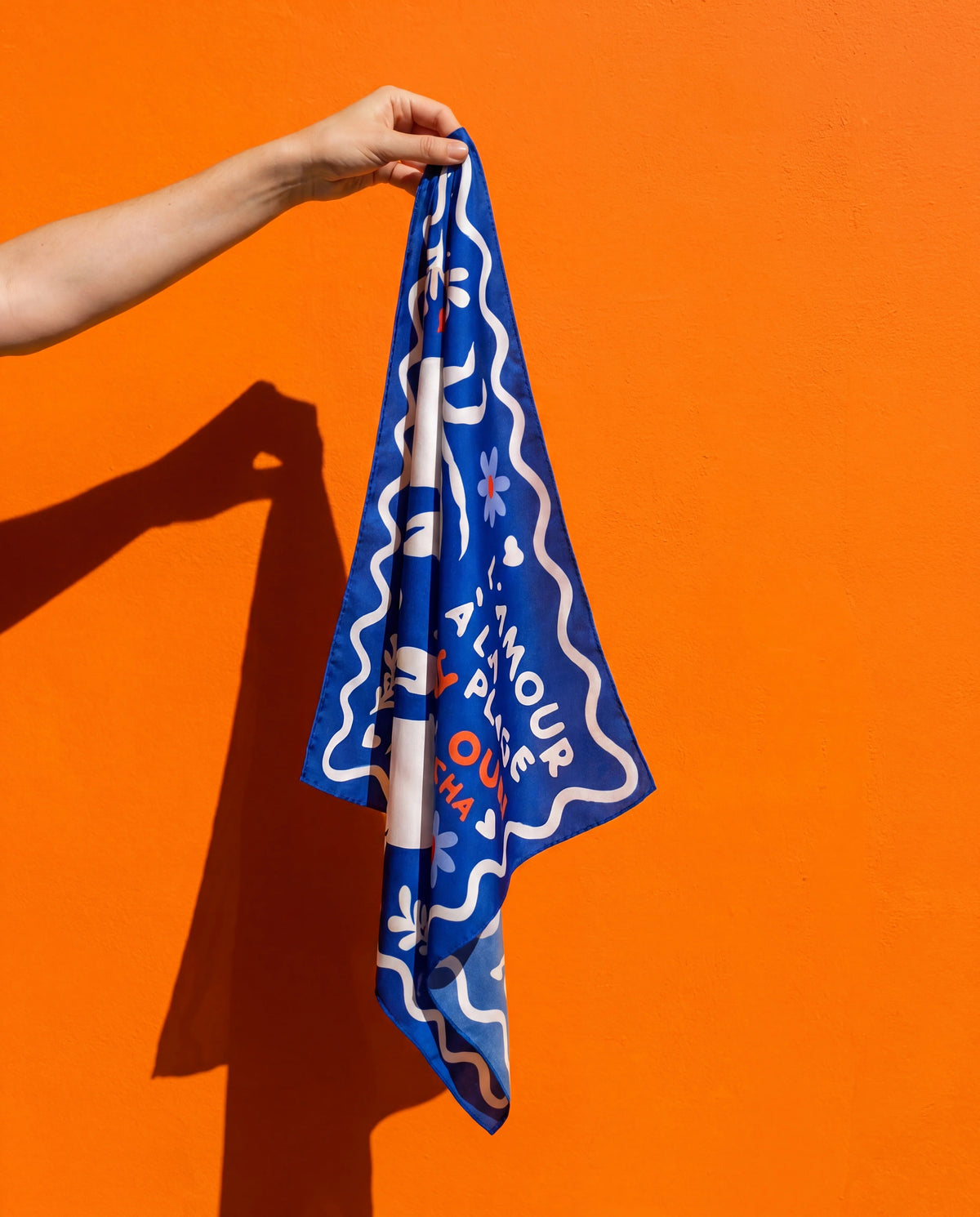 Foulard L'Amour à la Plage Soleil Mojito en satin bleu, motif graphique, tenu en main sur fond orange