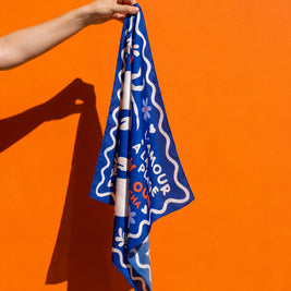 Foulard L'Amour à la Plage Soleil Mojito en satin bleu, motif graphique, tenu en main sur fond orange